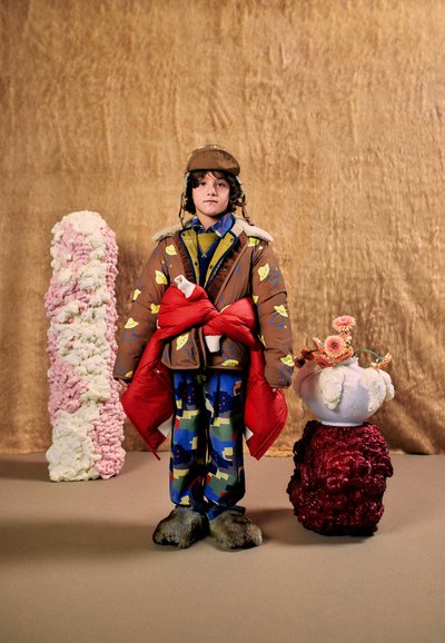 Un enfant mannequin porte une tenue colorée à motifs, une casquette marron et de grosses moufles rouges, entre des installations sculpturales texturées sur un décor de mode.