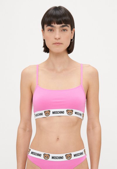 Rozā topiņš ar plānām siksniņām, kuram ir balta josla ar lāča logotipu un "MOSCHINO" tekstu. Atbilstoši rozā apakšbikses ar līdzīgu joslu.