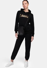 Black sweatshirt with gold "Lonsdale" logo, black jogger pants, and white sneakers. The model carries a small black crossbody bag.
