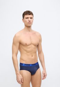 Άνδρας χωρίς πουκάμισο που στέκεται με το πρόσωπο προς τα εμπρός, φοράει μπλε σκούρα εσώρουχα Puma με μπλε μέση, πάνω σε απλό φωτεινό φόντο.