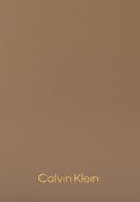 Peňaženka Calvin Klein v hnedej štruktúrovanej koži s vyrazeným logom v zlatej farbe na dolnom okraji. Povrch sa zdá byť hladký s jemným leskom.