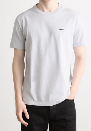 Homme portant un t-shirt gris clair uni à manches courtes avec un petit logo noir "BOSS" sur la poitrine, associé à un pantalon noir, debout devant un fond blanc.