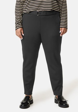 Personne portant un pantalon noir fuselé avec une ceinture, des mocassins noirs à franges, et une chemise à manches longues rayée noire et beige, debout sur un sol blanc.