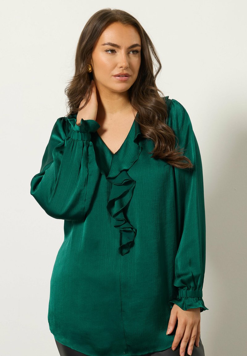 Smargdgrüne Bluse aus leichtem Stoff. Mit Rüschensaum am V-Ausschnitt, langen Ärmeln mit elastischen Bündchen und lockerer Passform.