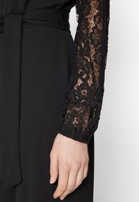 Svart byxdress med spetsärmar med blommönster. Slät tyg med knytbälte i midjan. Texturerad spets ger kontrast.