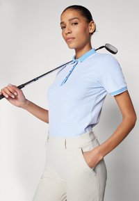 Jasnoniebieska koszulka polo z fakturowanym wzorem i kontrastowym kołnierzykiem, w połączeniu z beżowymi spodniami; model trzymający kij golfowy.