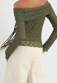 Schulterfreies Oberteil aus olivgrüner Spitze mit strukturiertem Design, langen Ärmeln und gekerbtem Saum. Kombiniert mit cremefarbenen, weit geschnittenen Hosen.