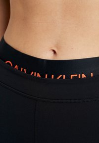 Leggings sportivi neri con una fascia in vita spessa e il logo "Calvin Klein" arancione. Il materiale sembra liscio e elastico.