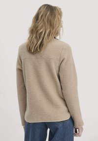 Beiger Fleece-Pullover mit Rundhalsausschnitt und langen Ärmeln. Mit einer Nahtdetails über den Rücken und einer lockeren Passform. Kombiniert mit blauen Jeans.