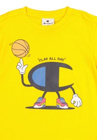 Camiseta de algodón amarilla con un personaje de dibujos animados equilibrando una pelota de baloncesto, con acentos en azul y negro y zapatillas coloridas. Texto: "JUEGA TODO EL DÍA."