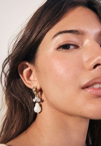 Gold hoop earring with a large white pearl design, embellished with clear crystals. Features smooth texture and rounded shapes.