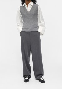 Gilet gris en tricot avec un col en V, superposé à une chemise blanche à boutons. Pantalons gris à jambe large et mocassins noirs avec des d�étails blancs.