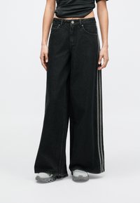 Person wearing wide-leg black jeans with side stripes, white and gray sneakers, and a cropped black top, standing against a plain background.