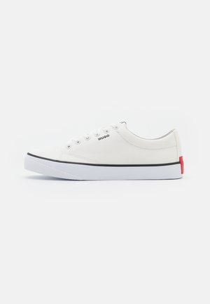 Hvid lavt snørehul lærreds-sneaker med hvide snørebånd, sort kant på sålen, rød detalje på hælen og "HUGO"-logo på siden.