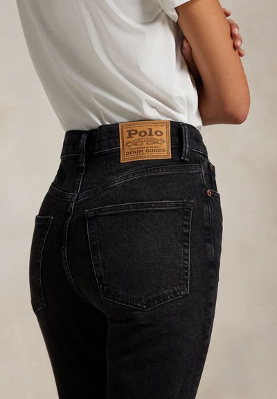 Persoon draagt zwarte high-waisted jeans met een bruine Polo Ralph Lauren leren patch op de tailleband, gecombineerd met een wit ingestopt overhemd.