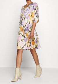 Blumenkleid mit V-Ausschnitt, das lila, orange und rosa Muster auf einem hellen Hintergrund zeigt. Kombiniert mit beigen High-Heels-Stiefeln.