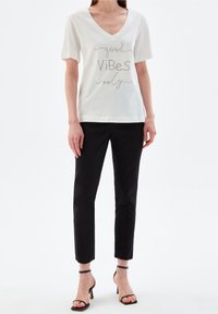 Bílý tričko s krátkým rukávem a výstřihem do V s dekorativním textem "dobré vibrace jen" ve stříbrné barvě, zkombinované s černými slim-fit kalhotami a podpatky.