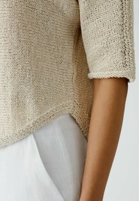 Beiger Strickpullover mit kurzen Ärmeln, der ein strukturiertes, lockeres Gewebe aufweist; kombiniert mit hellen, glatten Stoffhosen.