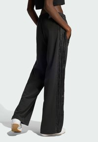 Black athletic pants with a relaxed fit, side pockets, and textured stripe accents down the legs, paired with white sneakers.