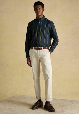 Mann steht mit Händen in den Taschen, trägt ein dunkel gestreiftes Hemd mit Knöpfen, beige Hose, brauen Gürtel, gemusterte Socken und braune Schuhe.