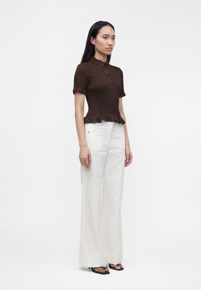 Femme portant un polo marron texturé à manches courtes, un pantalon blanc à jambes larges avec poches avant et des sandales noires ouvertes.