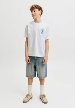 Nooruki poiss, kellel on lokkis juuksed, valge T-särk rohelise logoga, teksashoidised, valged sokid ja beežid kingad, seisab ja naeratab, näoga vasakule.