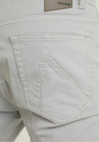 Grijze katoenen broek met een kenmerkend schuin achterzakdesign, zichtbare stiksels en een leren merkpatch. Gladde stoftextuur.