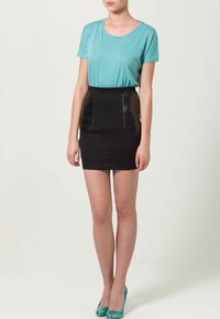 Un top turquoise a maniche corte è abbinato a una gonna nera con dettagli in ecopelle in vita. La modella indossa tacchi alte teala.