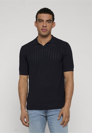 Mężczyzna ubrany w czarną dzianinową koszulkę polo z krótkim rękawem o fakturowanym wzorze oraz jasnoniebieskie dżinsy, stojący na tle jasnego, jednolitego tła.