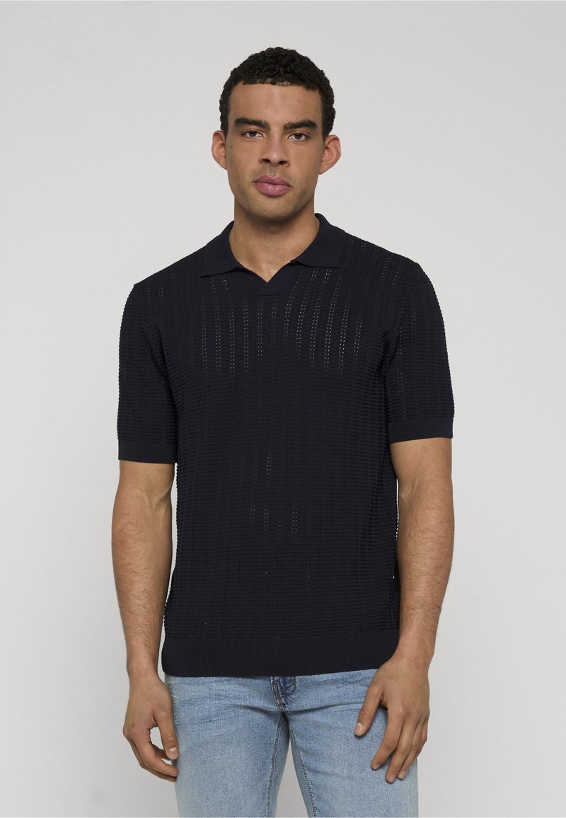 Homme portant un polo en maille noir à manches courtes avec un motif texturé et un jean bleu clair, debout devant un fond clair uni.