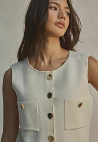 Gilet senza maniche di colore bianco sporco con due tasche frontali, bottoni dorati e tessuto testurizzato. Collo rotondo con finitura liscia.