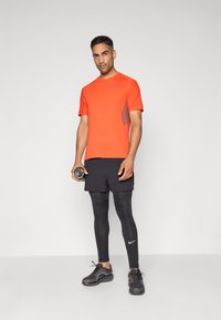 Mand i lys orange kortærmet skjorte, sorte shorts over sorte leggings, holder en vandflaske og står mod en simpel lys baggrund.