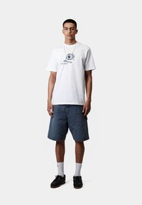 Camiseta blanca de algodón con un estampado gráfico azul, combinada con shorts de mezclilla azules, calcetas blancas y zapatos negros. Diseño simple.