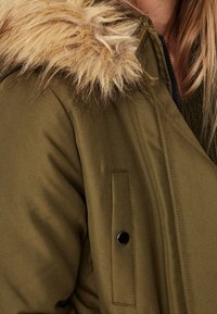 Olive green jacket featuring a faux fur hood trim, a zip closure, and a side pocket with a black snap fastener. Smooth, textured fabric.