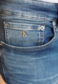 Jeans vaqueros Calvin Klein con un acabado azul desteñido, textura desgastada, diseño clásico de cinco bolsillos y un logo bordado en amarillo en el bolsillo trasero.