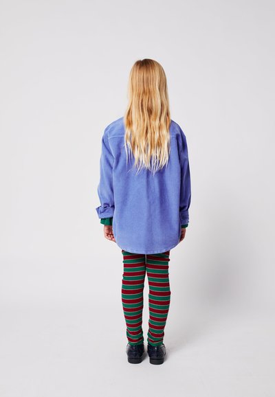 Camicia oversize azzurro chiaro con maniche lunghe, abbinata a leggings a righe rosse e verdi. Tessuto morbido, design casual. Scarpe nere.