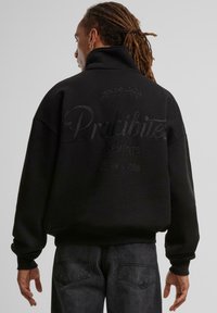 Sorth genser med høy krage, oversized passform, med ton-i-ton broderi på baksiden. Laget av mykt stoff med ribbestrikket mansjetter.