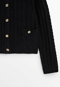 Musta teksturoitu neulecardigan, jossa on pitkät hihat, kultaiset koristepainikkeet etuosassa ja pienessä rintataskussa.