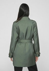 Zielony płaszcz typu trench, wykonany z gładkiego materiału, z paskiem, zapięciem na guziki przy mankietach oraz rozcięciem z tyłu, z klasycznym kołnierzem i dopasowanym krojem.