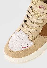 Beige og hvite Nike-sko med perforert tåboks, beige snøringer, brun sidepanel og liten rød Nike Swoosh på fronten.