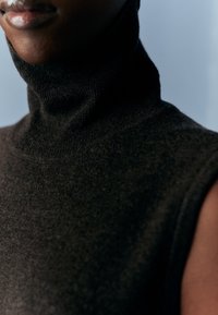 Gros plan d'une personne portant un pull à col roulé sans manches foncé sur un fond gris atténué.