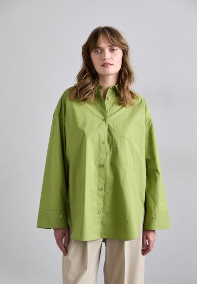 Chemise oversized vert clair avec un devant à boutons, une petite poche et des manches longues. Portée avec un pantalon ample de couleur claire.