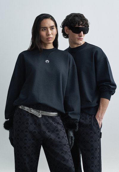 Due figure indossano felpe nere abbinate con un emblema circolare e pantaloni neri con motivo. I pantaloni presentano un design con un logo sottile.