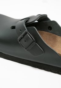 Sandale en cuir noir avec une fermeture à boucle, une semelle intérieure en liège et une semelle extérieure en caoutchouc texturé. Forme arrondie avec un design à dos ouvert.