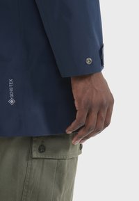 Mornarska GORE-TEX jakna s dugmetima na manžetama, koja sadrži vidljivi logo. Ruka se odmara na maslinasto zelenim cargo hlačama s dugmetima.