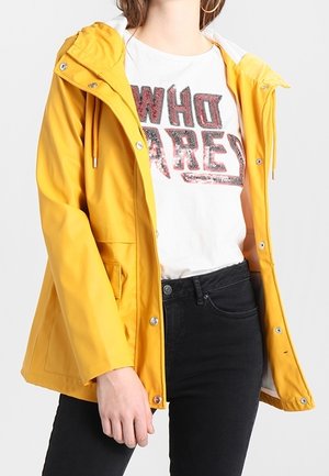 Personne portant une veste à capuche jaune sur un T-shirt blanc avec le texte "WHO ARE" et un jean noir, debout devant un fond uni.
