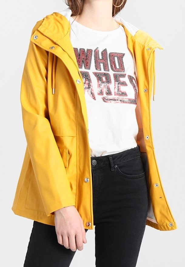 Personne portant une veste à capuche jaune sur un T-shirt blanc avec le texte "WHO ARE" et un jean noir, debout devant un fond uni.