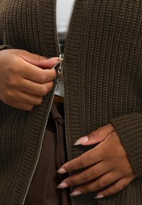 Brun zip-up cardigan med ribbad textur, silverdragkedja och vida ärmar, lager på lager över en vit topp och bruna byxor.