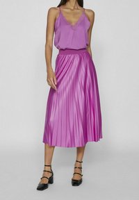 Camisola di raso viola con pizzo, abbinata a una gonna rosa plissettata di lunghezza al ginocchio. Il modello indossa scarpe con tacco blocco e cinghie alla caviglia.