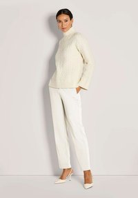 Vrouw gekleed in een crèmekleurige kabelgebreide coltrui, witte op maat gemaakte broek en beige slingback hakken, staand tegen een lichtgrijze achtergrond.